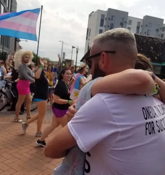 An embrace at a Pride gathering, with trans flags in the crowd.