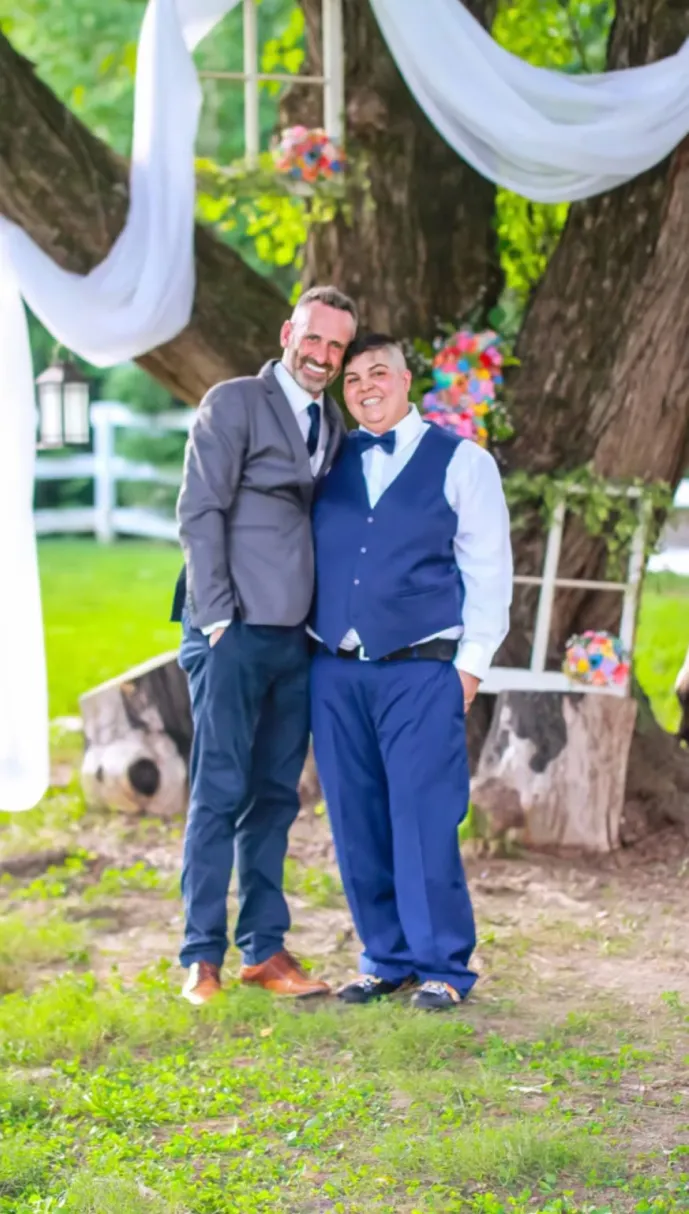Two smiling people in wedding suits beneath a tree decorated with white fabric and rainbow flowers.