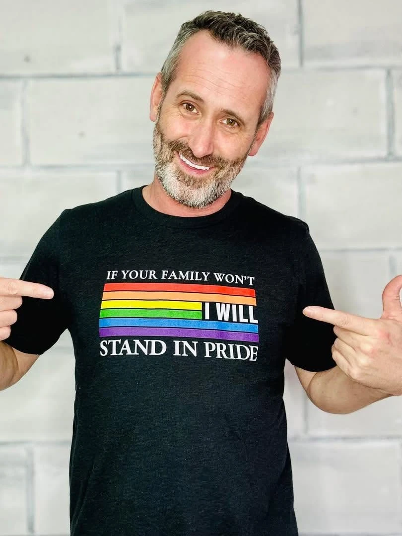 Daniel Blevins smiling and pointing to his Stand In Pride shirt, which reads: If your family won't, I will.